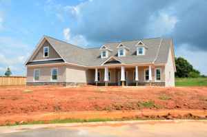 A new residential house under construction, featuring a light-colored exterior, multiple gables, and a large front porch, set against a backdrop of blue sky and green landscape.